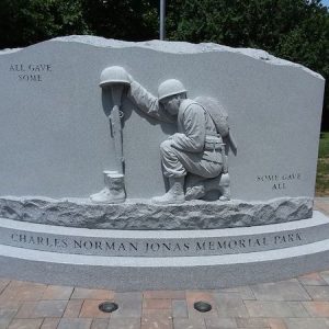 The Charles Norman Jonas Memorial Park consists of a gray granite upright. It features a sculpture of a kneeling soldier over a grave marked by boots, a rifle, and a helmet. The memorial has the phrase "All Gave Some, Some Gave All."