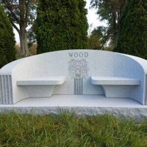 The Wood Family Estate Bench Memorial is crafted from gray Rock of Ages granite. It is formed as a dual bench configuration with a central upright featuring a carving of the family name and floral arrangement. This memorial is suitable for use with traditional interment or cremation.