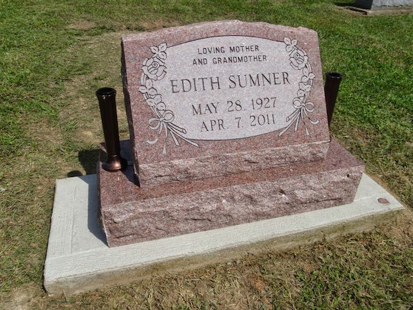 This Single Slant Marker on a Base honoring Sumner is crafted from pink granite and is flanked by two vases. It features engraved floral accents around an oval memorial information area. This memorial is suitable for use with cremation or traditional interment.
