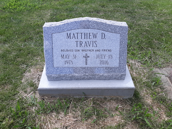 This Single Slant Marker honoring Travis is crafted from gray granite. It features an engraved cross. It is appropriate for use with cremation or traditional interment.