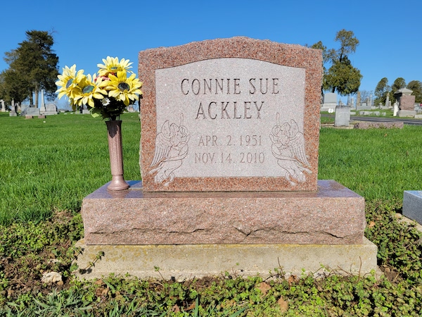 This Single Upright honoring Ackley is crafted from red granite and is flanked with a vase. It features carvings of two angels on either side of the memorial information. This memorial is suitable for use with traditional interment or cremation.