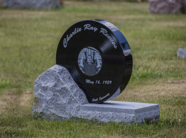 This Custom Single Upright with Record shaped stone crafted from polished black granite is set into a rough hewn gray granite base. The record portion of the memorial features a laser engraved image of the decedent acting as a disc jockey playing music. This unique memorial is appropriate for use with cremation or traditional interment.