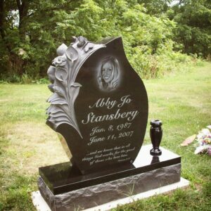 This Custom Single Upright with Lilies carved into the flame shaped stone is crafted from polished black granite. The monument features a laser engraving of the decedent and is flanked with a single vase. This type of monument is suitable for use with cremation or traditional interment.