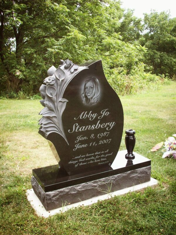 This Custom Single Upright with Lilies carved into the flame shaped stone is crafted from polished black granite. The monument features a laser engraving of the decedent and is flanked with a single vase. This type of monument is suitable for use with cremation or traditional interment.