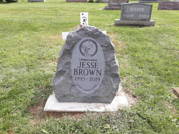 This single boulder memorial is crafted from a piece of rough brown granite and features a carved image of a tree in an oval memorial plate on the stone. This marker is suitable for traditional interment or cremation.