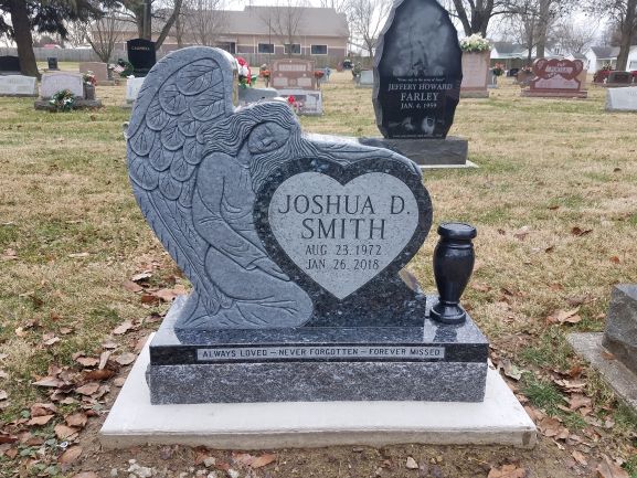 This style of memorial is a. Custom Single Upright with Angel. Crafted from blue marble, the marker has the shape of an angel watching over a heart shaped portion of the stone. It is a custom carving on the memorial and is appropriate for traditional interment or cremation.