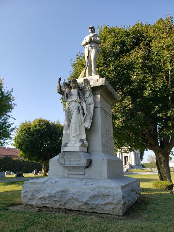 The Historic George Harper War Memorial is crafted from gray granite. It is a multi-part memorial featuring a large 3D carving of Harper wearing a uniform and holding a rifle at the top of an ornately carved four-sided pedestal with carvings memorializing his deeds. A large 3D angel with wings sculpted from white granite stands in front of the pedestal.