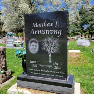 This Single Upright with Skateboard is crafted from polished black granite and is flanked by a vase. The monument features a photo realistic laser engraved image of the decedent and a carved skateboard and tree. This memorial is appropriate for use with cremation or traditional interment.