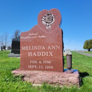 This Custom Single Upright with Rose in Heart is crafted from red granite and is flanked with a vase. The stone is in the outline of a heart and features a carved single rose in that area. This monument is appropriate for use in traditional interment or cremation.