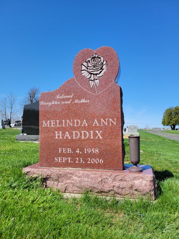 This Custom Single Upright with Rose in Heart is crafted from red granite and is flanked with a vase. The stone is in the outline of a heart and features a carved single rose in that area. This monument is appropriate for use in traditional interment or cremation.