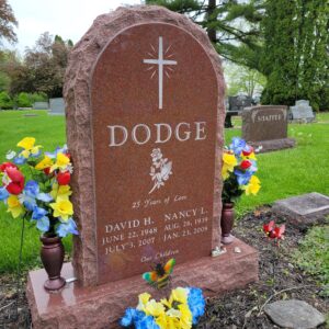 This Custom Companion Upright honoring Dodge is crafted from polished red granite and is flanked by two vases. It features engravings of a cross and rose with hands in prayer. This memorial is suitable for traditional interment or cremation.
