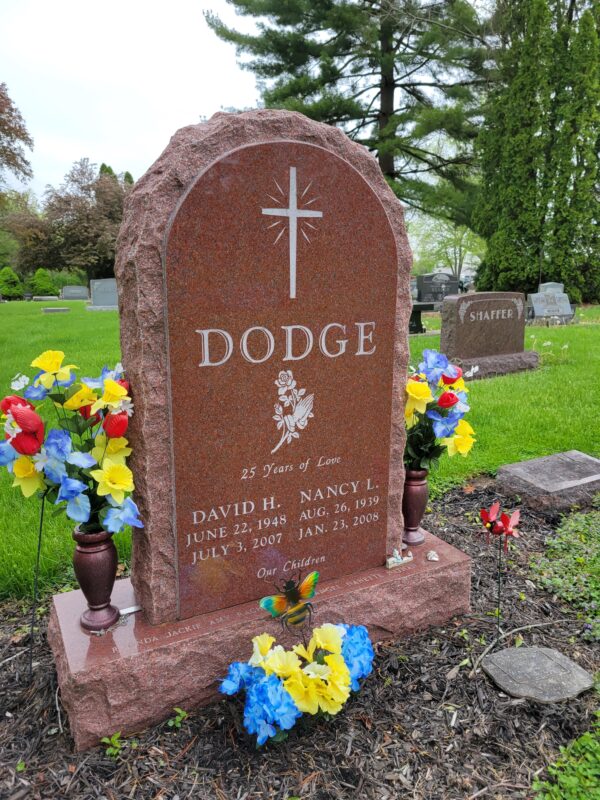 This Custom Companion Upright honoring Dodge is crafted from polished red granite and is flanked by two vases. It features engravings of a cross and rose with hands in prayer. This memorial is suitable for traditional interment or cremation.