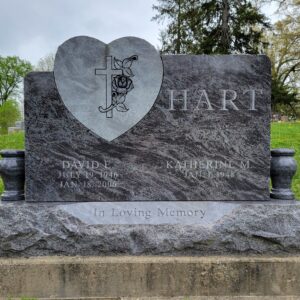 This Custom Companion Upright honoring Hart is crafted from polished blue granite and is flanked by two vases. It features a stone shaped like a heart that is filled with an engraved cross with roses. This memorial is suitable for traditional interment or cremation.
