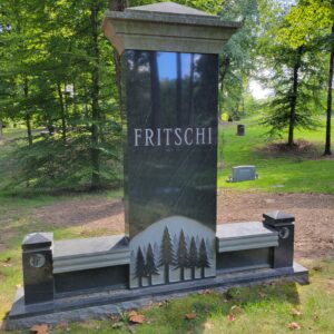 The Fritschi Family Estate Memorial is crafted from polished black Rock of Ages granite. It features a large central upright flanked by twin benches. The upright base has a carving of a stand of pine trees. This memorial is appropriate for use with traditional interment or cremation.