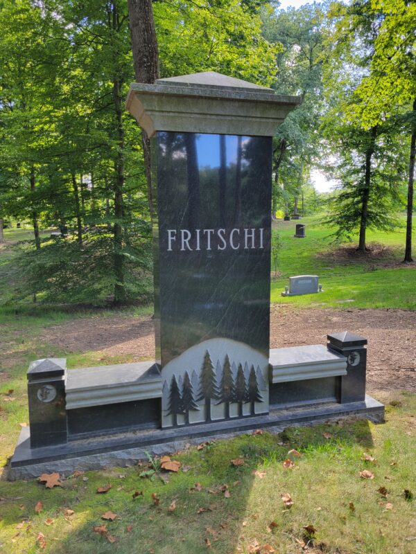 The Fritschi Family Estate Memorial is crafted from polished black Rock of Ages granite. It features a large central upright flanked by twin benches. The upright base has a carving of a stand of pine trees. This memorial is appropriate for use with traditional interment or cremation.