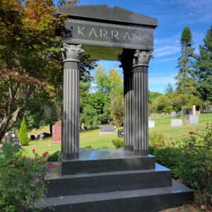 The Karras Family Estate Memorial is crafted from polished and rough black granite. It consists of a platform supporting four Greek style columns and a Greek temple style roof. The roof area has the Karras family name engraved and features a geometric scrollwork pattern. This memorial is appropriate for use with cremation or traditional interment.