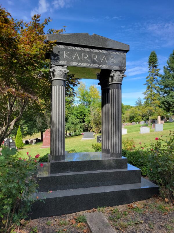 The Karras Family Estate Memorial is crafted from polished and rough black granite. It consists of a platform supporting four Greek style columns and a Greek temple style roof. The roof area has the Karras family name engraved and features a geometric scrollwork pattern. This memorial is appropriate for use with cremation or traditional interment.