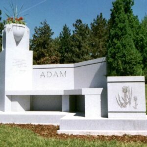 The Adam Family Estate Memorial is crafted from white granite. It consists of multiple components including a tower serving as a bas for a large vase, a wide upright with the family named engraved prominently, a bench, and a cube with a floral carving. This memorial is appropriate for use with cremation or traditional interment.