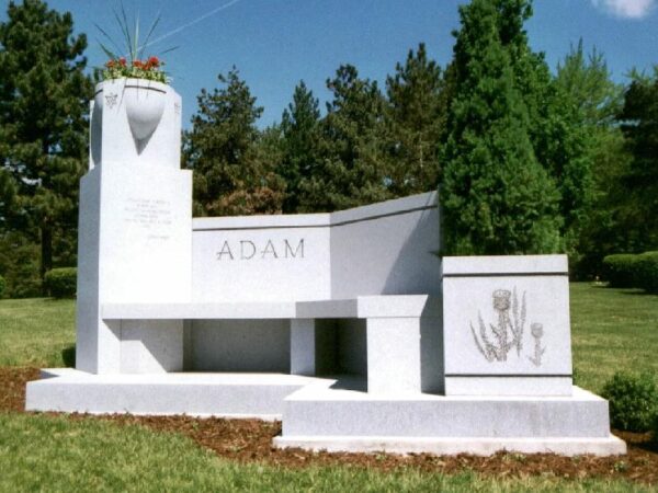 The Adam Family Estate Memorial is crafted from white granite. It consists of multiple components including a tower serving as a bas for a large vase, a wide upright with the family named engraved prominently, a bench, and a cube with a floral carving. This memorial is appropriate for use with cremation or traditional interment.