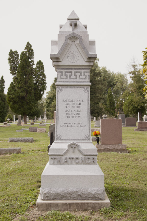The Thatcher Family Estate Memorial is crafted from gray granite and is in the shape of a four sided tower upright. It features an engraved clock near the top, an ornate roof structure, and several stylized capital letter T shapes throughout. This memorial is appropriate for use with cremation or traditional interment.