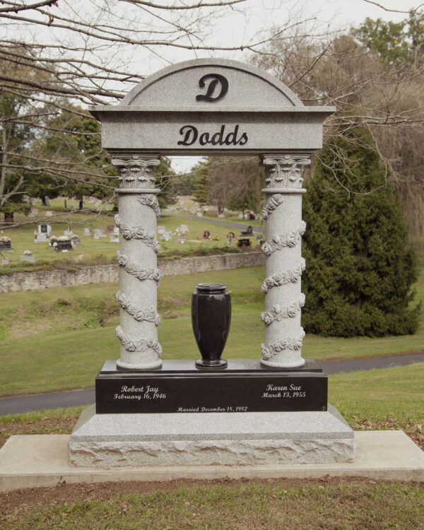 This Custom Companion Upright with Columns is crafted from gray and polished black granite. It features two columns with floral relief climbing toward a capstone with the family name. The columns are set on a polished black granite base with a vase and the memorialization details for the decedents. This memorial is appropriate for use with traditional interment or cremation.
