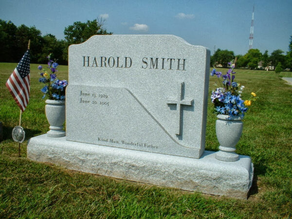 This Custom Single Upright with Two Vases is crafted from gray granite and is shaped with multiple levels of relief. It features a carving of a cross extending from the face of the stone. This monument is appropriate for use with traditional interment or cremation.