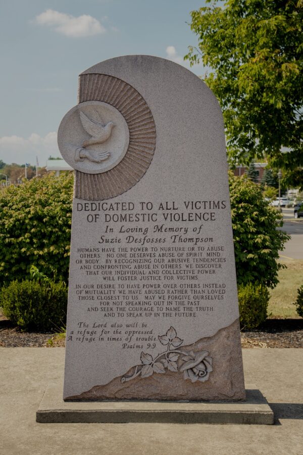 The Warren County Domestic Violence Memorial is crafted from pink granite and is the form of a sculpted upright. It features a carving of a sun with a dove and hand on the top with the stone sharing the form of the sun. The rest of the monument has engraved memorial information along with a single rose.