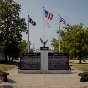 The Jeffersonville Veterans Memorial is crafted from gray and polished black granite. It consists of three uprights, one central gray granite upright supports a polished black pedestal with an eagle sculpture resting on it. The flanking polished black uprights contain the memorial information of the veterans honored with the memorial. There are three flag poles with the United States, State of Ohio, and POW/MIA flags displayed. You can see this memorial in person in Jeffersonville, Ohio.