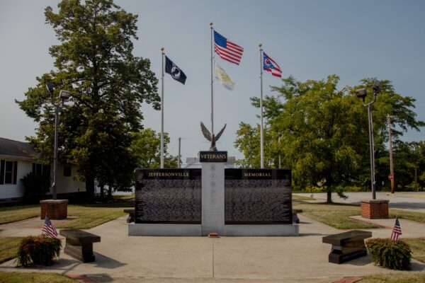 The Jeffersonville Veterans Memorial is crafted from gray and polished black granite. It consists of three uprights, one central gray granite upright supports a polished black pedestal with an eagle sculpture resting on it. The flanking polished black uprights contain the memorial information of the veterans honored with the memorial. There are three flag poles with the United States, State of Ohio, and POW/MIA flags displayed. You can see this memorial in person in Jeffersonville, Ohio.